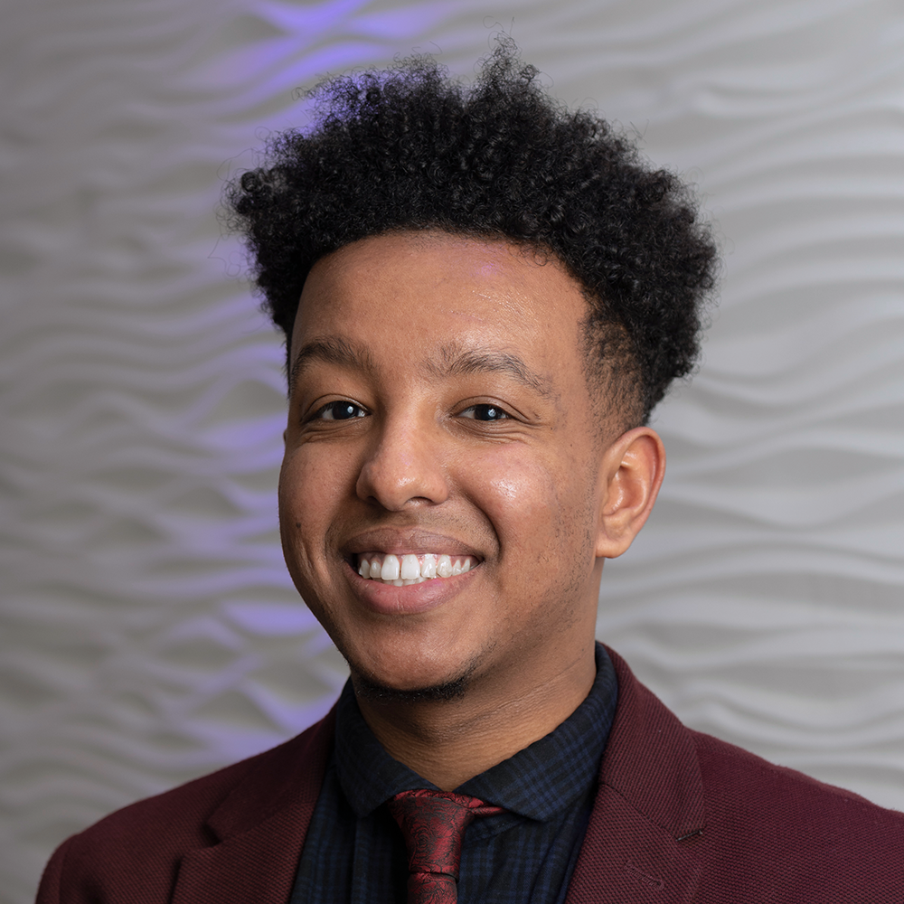 Manny Dinssa, founder of Plainly Innovate, smiling in a deep red blazer and patterned tie